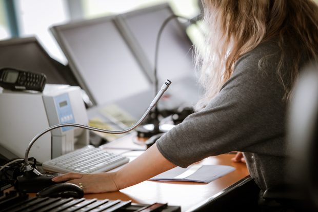 Profile view of woman air traffic controller seated at a desk on which there are 2 computers and a thin bendy microphone.