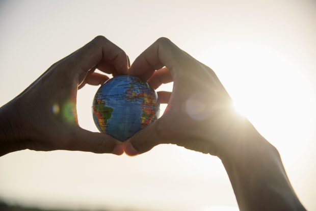 Pair of hands in aheart shape, holding a small Earth globe with sunshine coming in from behind the right hand.