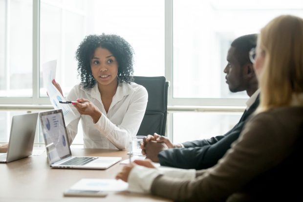 African businesswoman, sitting at a table, holding a report and taling to 2 colleagues.