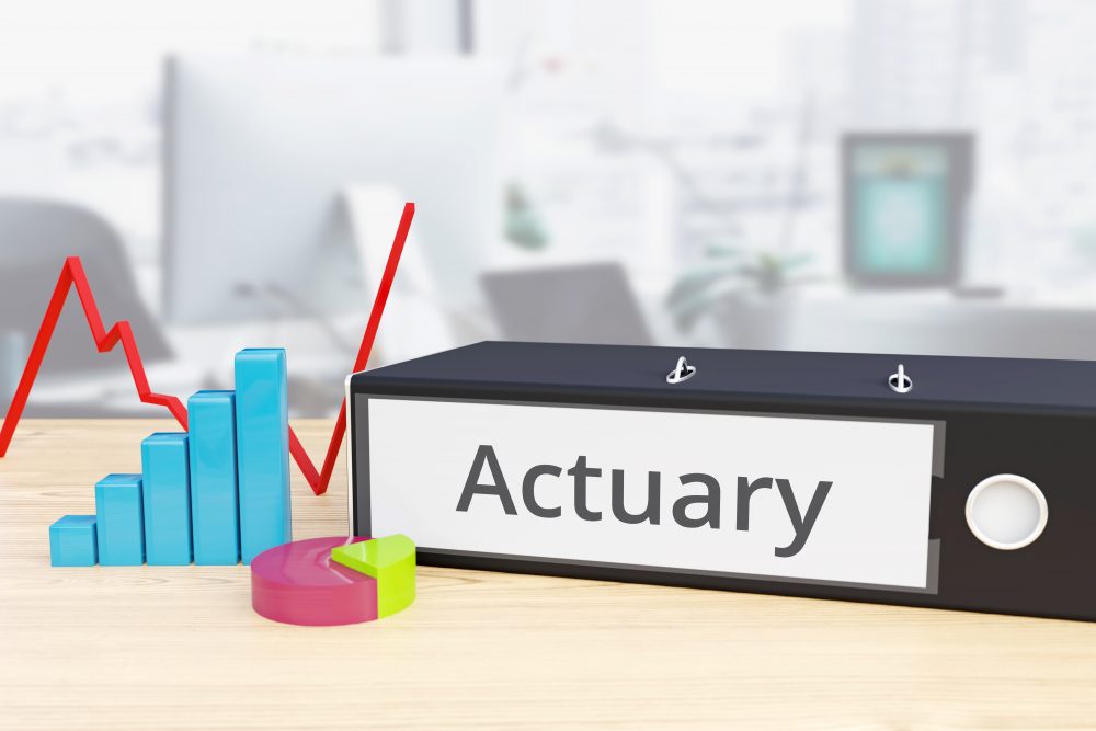 Lever arch file with the word actuary on its spine, on a table next to representations of a pie chart, graph and bar chart.