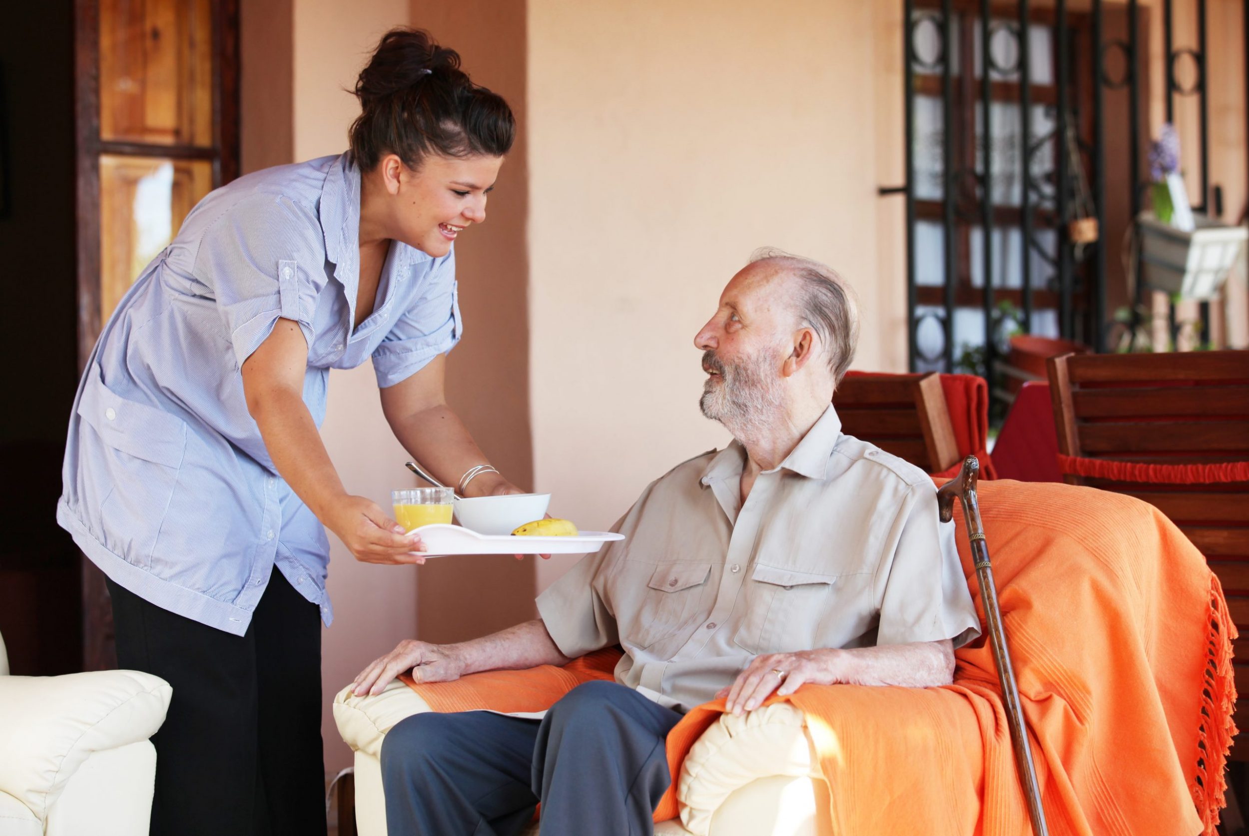 Female care home worker giving tray of food to seated care home resident.