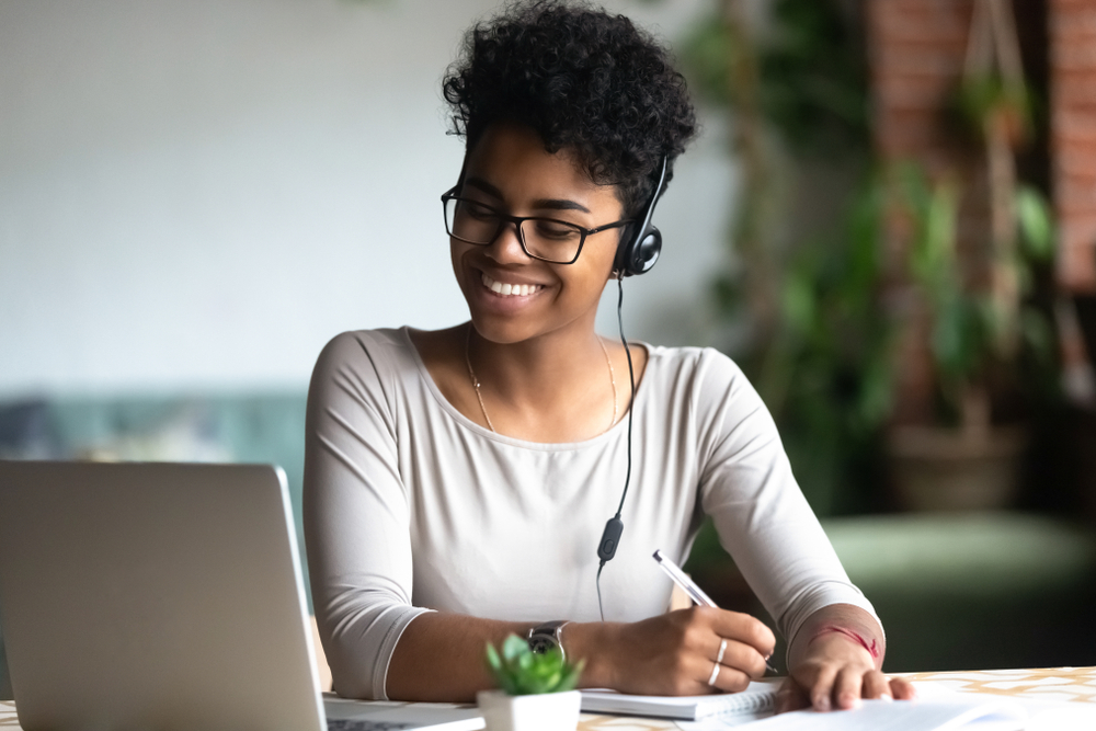 Smiling woman wearing headphones, looking at her laptop and writing on a notepad.