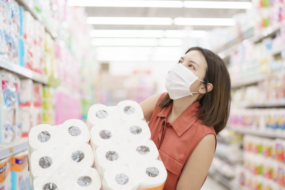 Woman in a supermarket, wearing face mask, carries 2 large packages of toilet rolls.