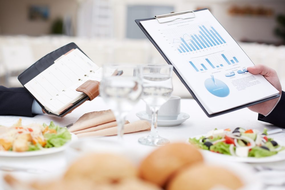 Dining table with salads, bread and glasses of water. One male hand holding a leather diary planner and another man's hand showing graphs on paper attached to a clipboard.