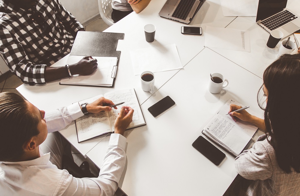 Image shows colleagues sitting at a white table, taking notes with notebooks and laptops. There are a few cups of coffee and phones on the table.