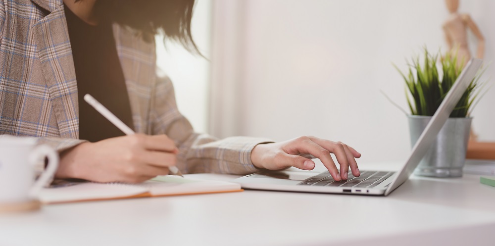 Image shows woman on laptop, with a notebook and pen.