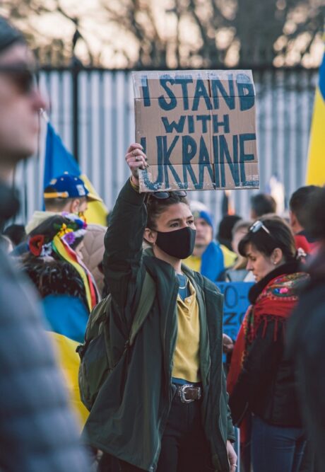 Woman wearing a face mask over her nose and mouth stands in a crowd of people. She is holding up a cardboard sign on which is written 'I stand with Ukraine'.