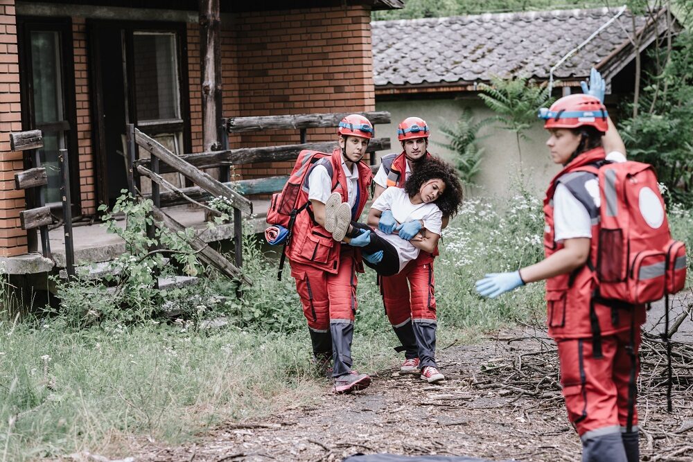 Two rescue workers in red clothing and red helmets carry and injured woman who has blood on her face. Anotehr rescue worker stands nearby raising her right hand as if to wave them on.