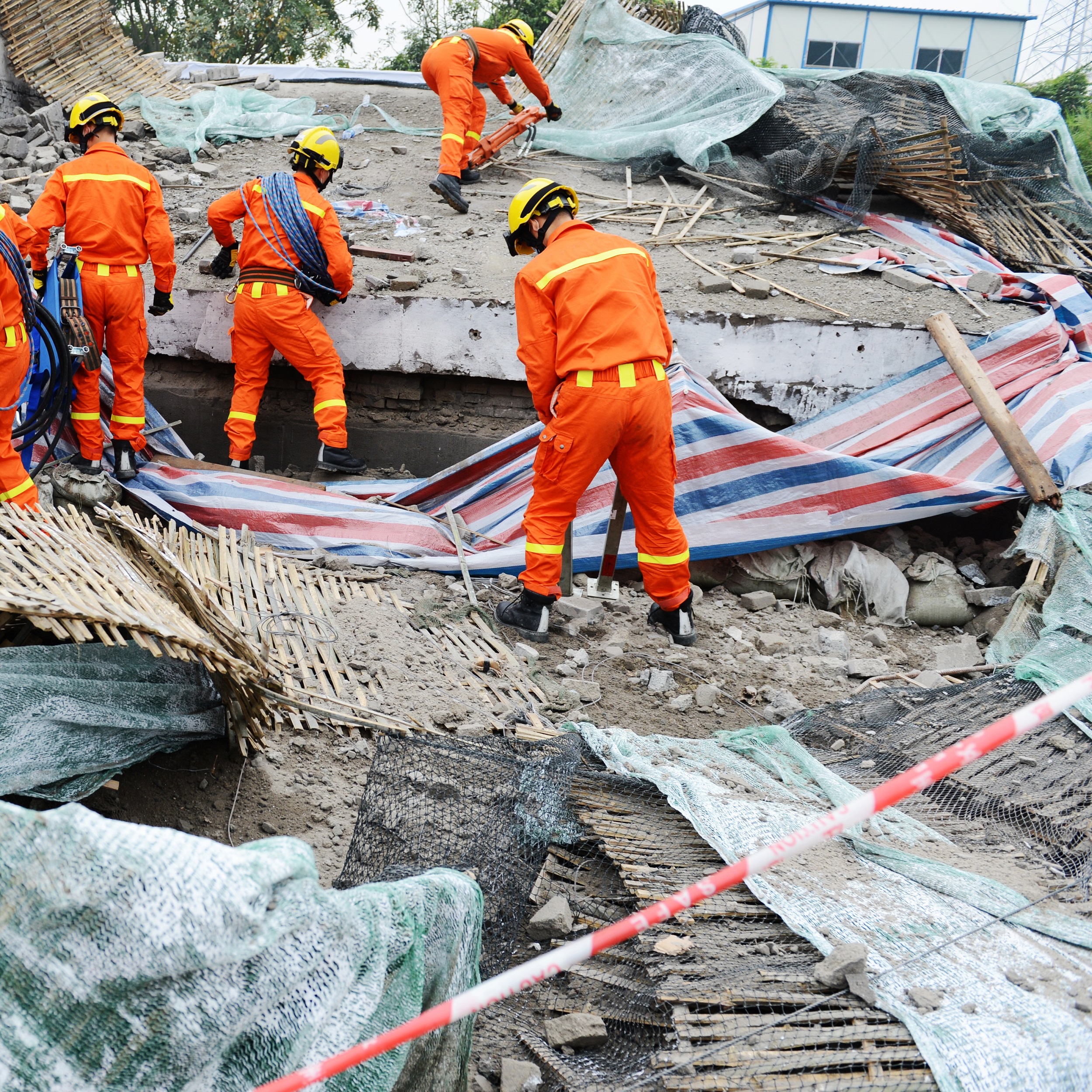 Rescue workers in bright orange clothing and yellow hardhats are seen climbing over the ruits of a building as they search for survivors after a disaster.