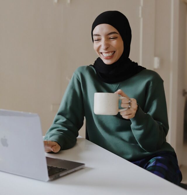 Smiling woman in hijab holding a mug in her left hand and sitting in front of a laptop.