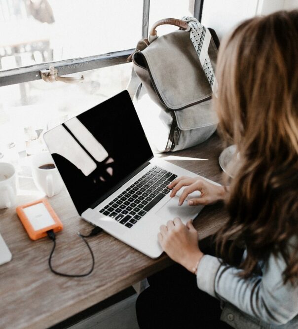 Rear view of woman with long brown hair who is working on a laptop on a desk facing a window. On the table is her rucksack bag, several coffee cups and her laptop is connected to an orange coloured charger.