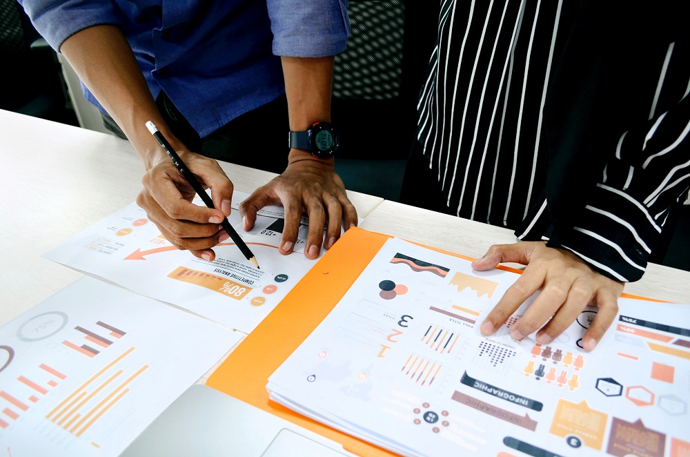 The hands of 2 people - one holding a pencil - are resting on images of data information on a desk.