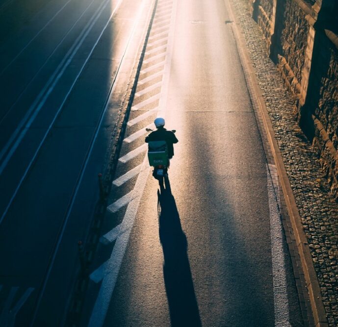 Aerial view of a person riding a moped on an empty street. The moped is heading towards sunlight further up the road.