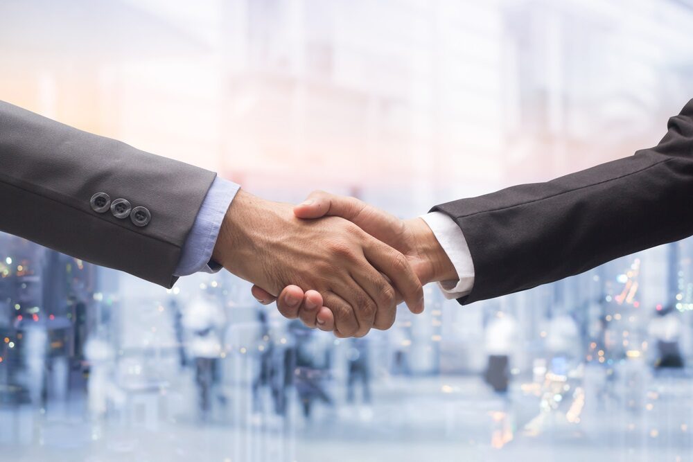 Image of a handshake of 2 white men wearing suit jackets. The image shows the suit sleeves, and hands.