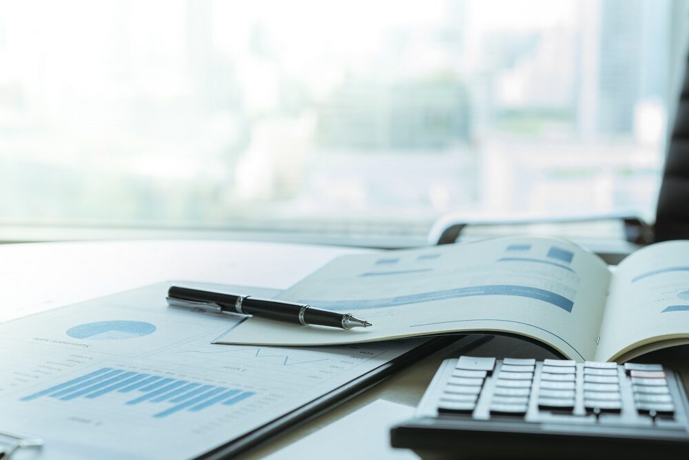 On a desk, there's a page of bar graphs is attached to a clipboard, a book showing corporate information and a calculator.