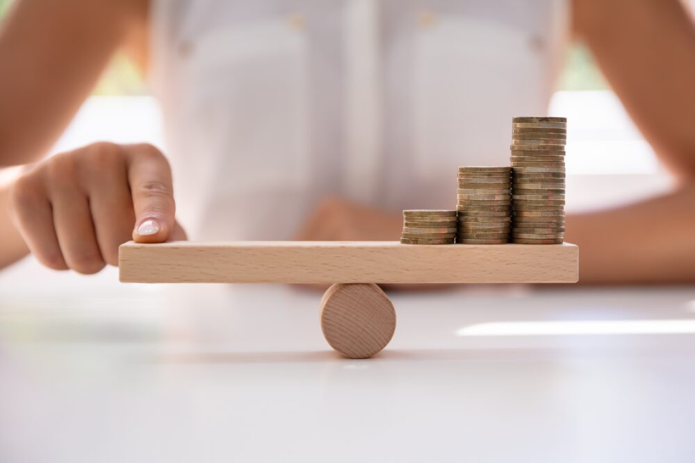 Person with their finger on a small wooden balance. On the other side is a stack of coins. The person is in the background of the picture and their image is blurred.