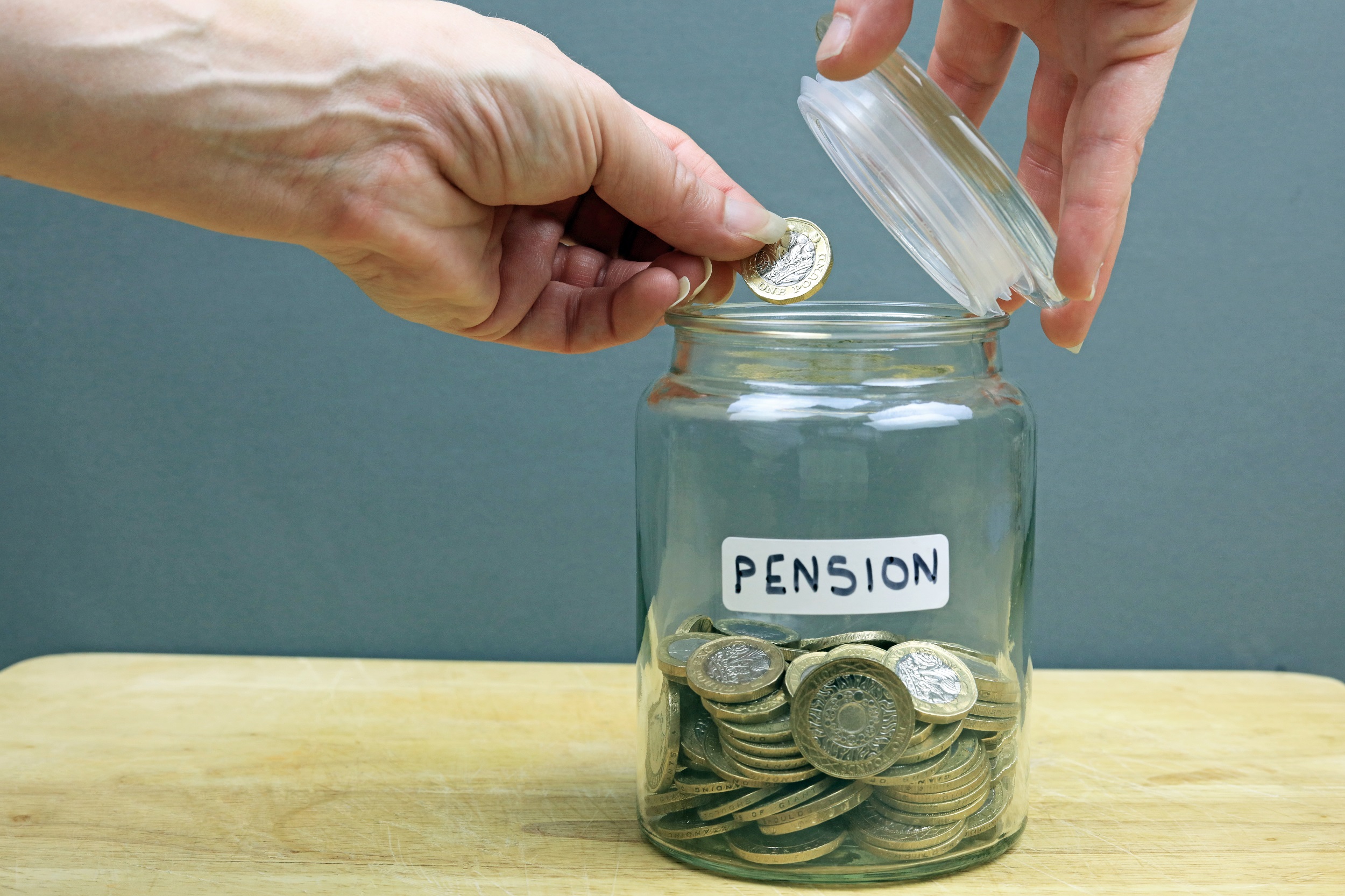 Close up of a person's hands as they place a pound coin into a glass jar which is half full of pound coins. On the jar there is a label with the word 'Pension' written on it.