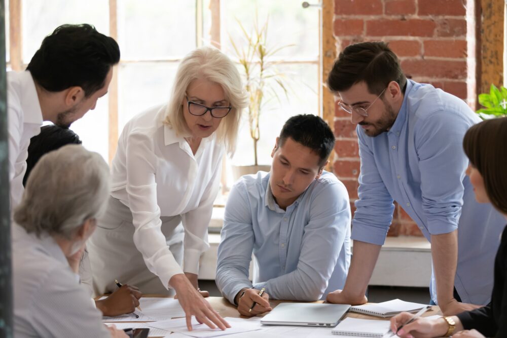 Group of people in a meeting around a table. Some are sitting and some are standing. A woman has her hand on paper on the table and everyone is looking down at what she is talking about.