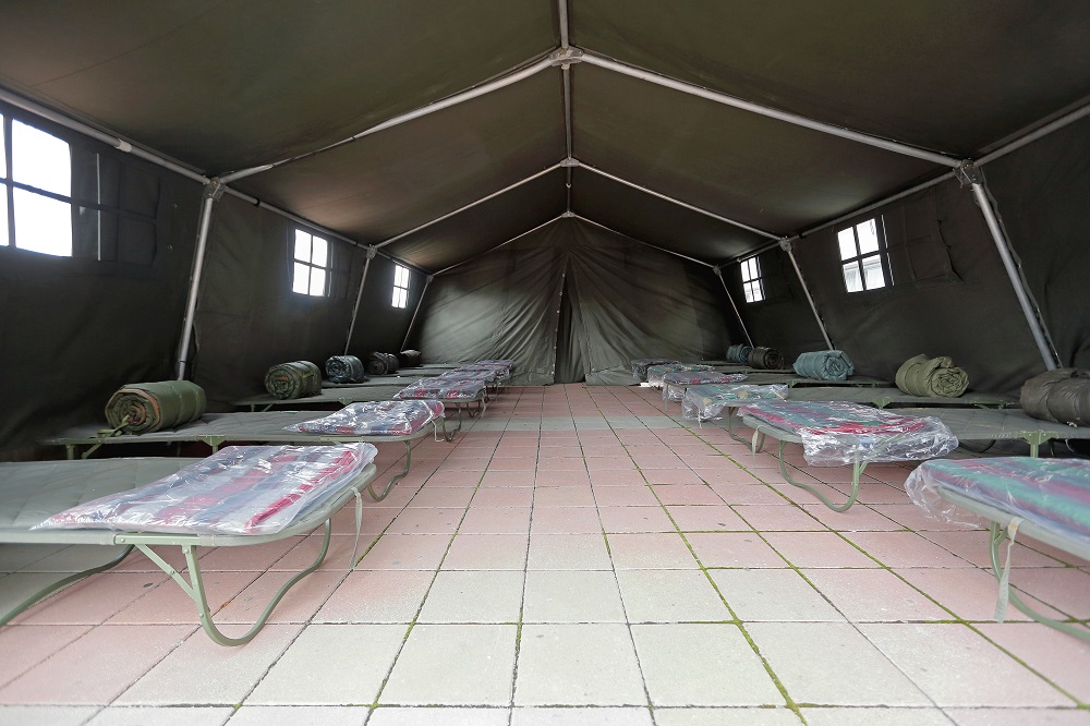 Two rows of camp beds in a large tent.