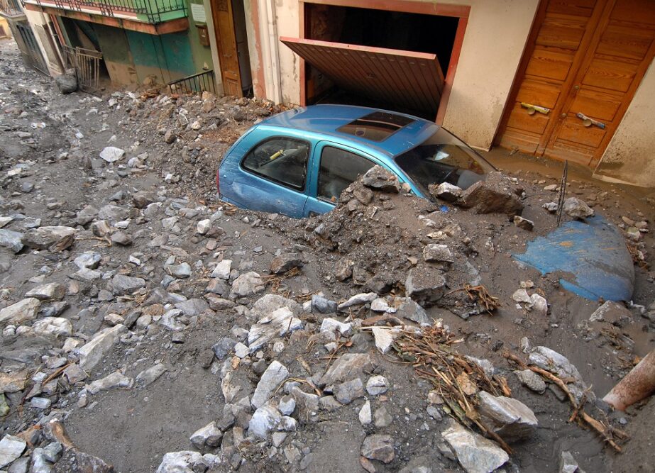 Blue car partially submerged in rock, sand and other debris from a landslide. The car is parked alongside a broken garage door with a front door next to it.