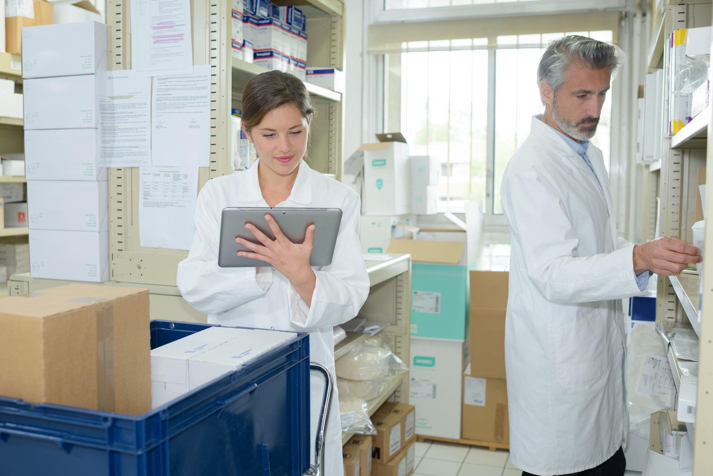 A man and a woman both wearing white coats are in a room with metal shelving. The woman is looking at an iPad and the man is checking stock on the shelves. The stock is all medical supplies in boxes.