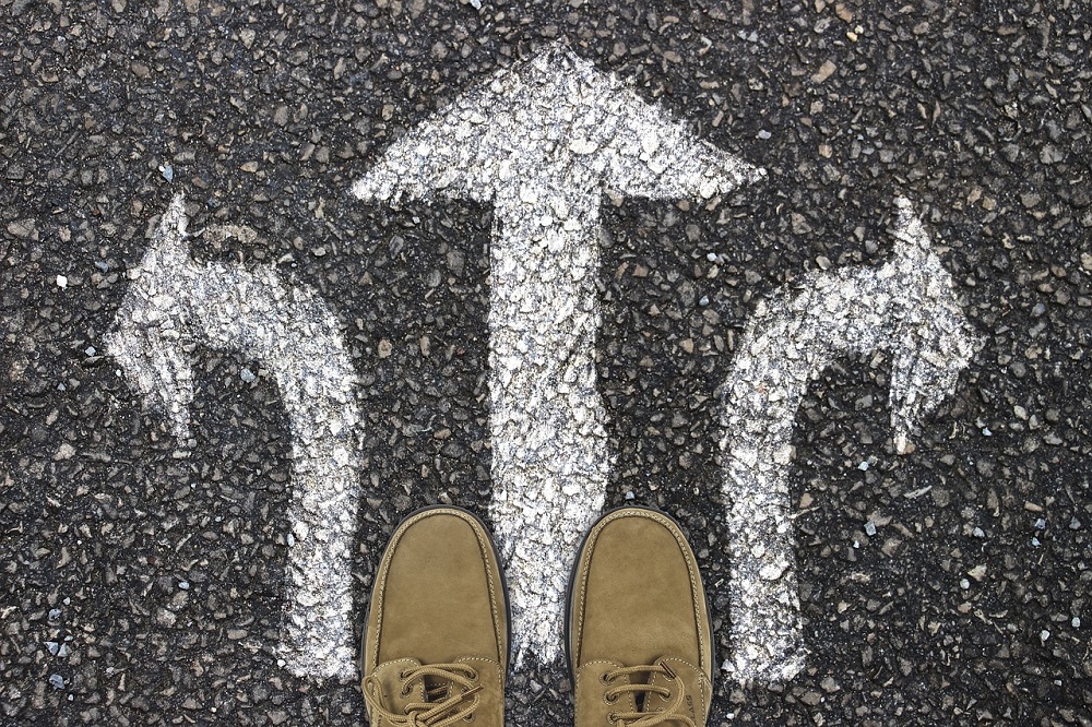 View from above of men's tan coloured lace up shoes standing on black tarmac where there are 3 arrows pointing ahead, to the left and to the right.