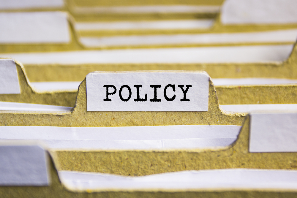 Close up view of cardboard filing dividers. One has a white tag, front and centre of the image, and has the word POLICY in capital letters typed on the tab.