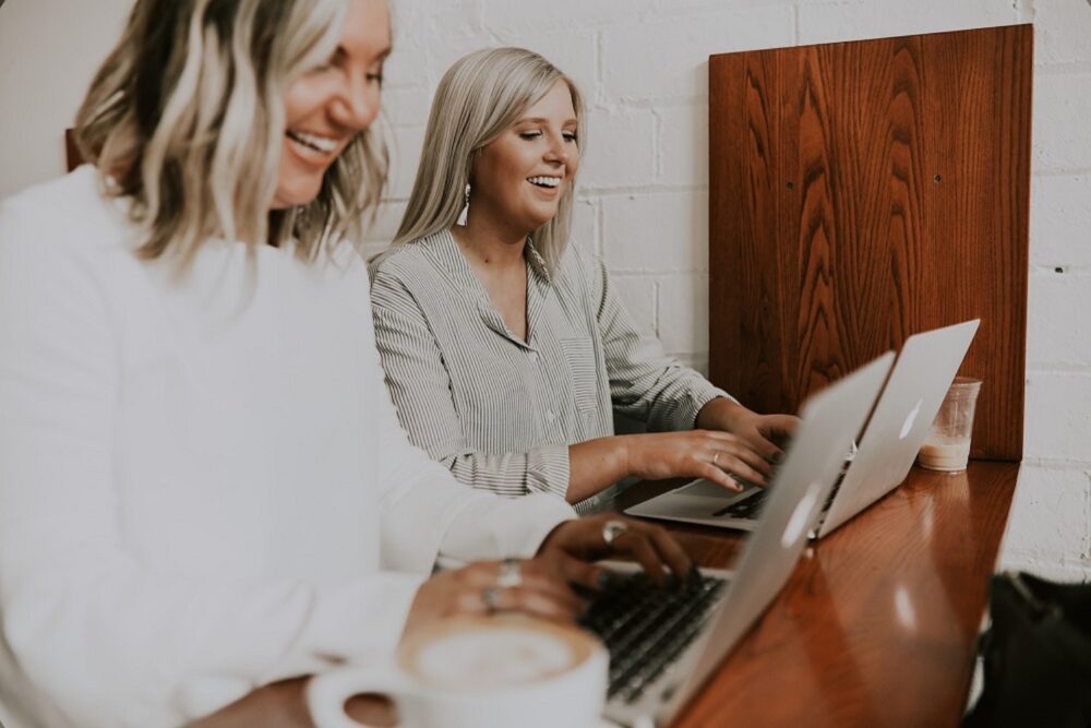 Two smiling women sitting side-by-side, working on their laptops.