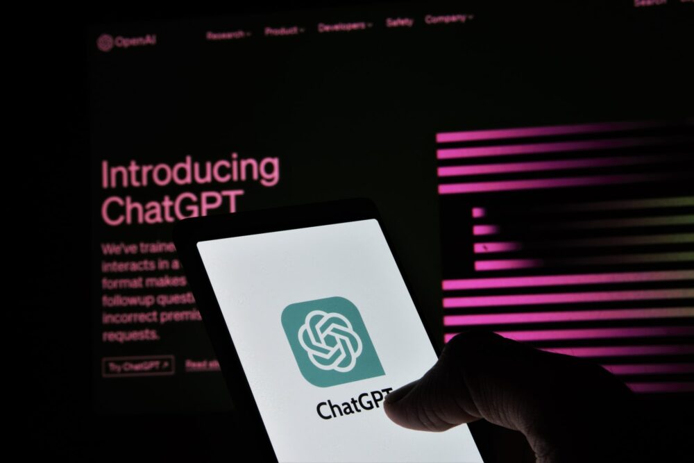 Hand holding a phone. The screen on the phone shows a white background and the words ChatGPT, along with a green logo. The phone is against a darkened laptop screen on which is written 'Introducing ChatGPT.'