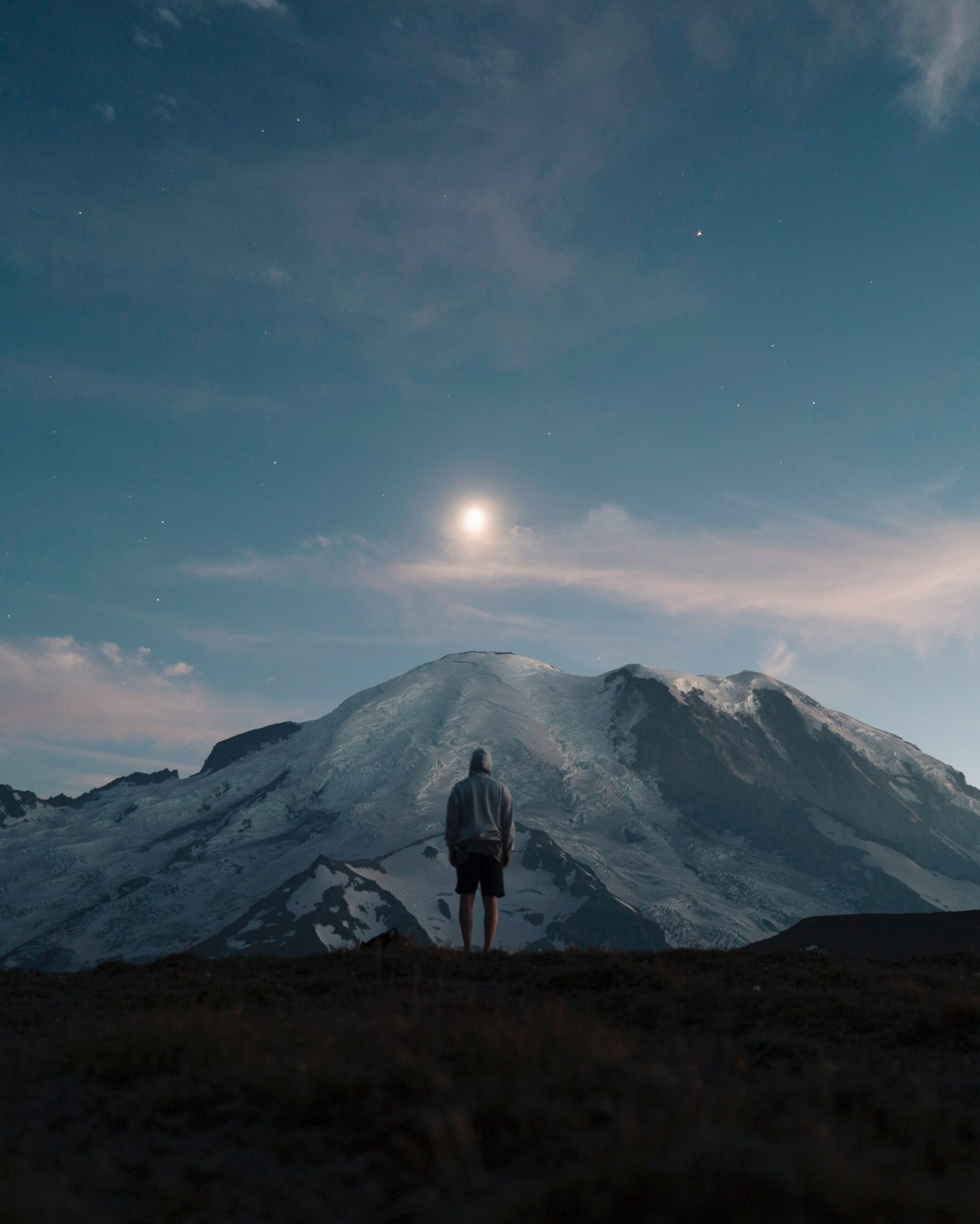Man standing looking at a mountain which is covered in snow. The image is taken in the early evening as the sky is a darker blue and some stars are visible, along with the moon.