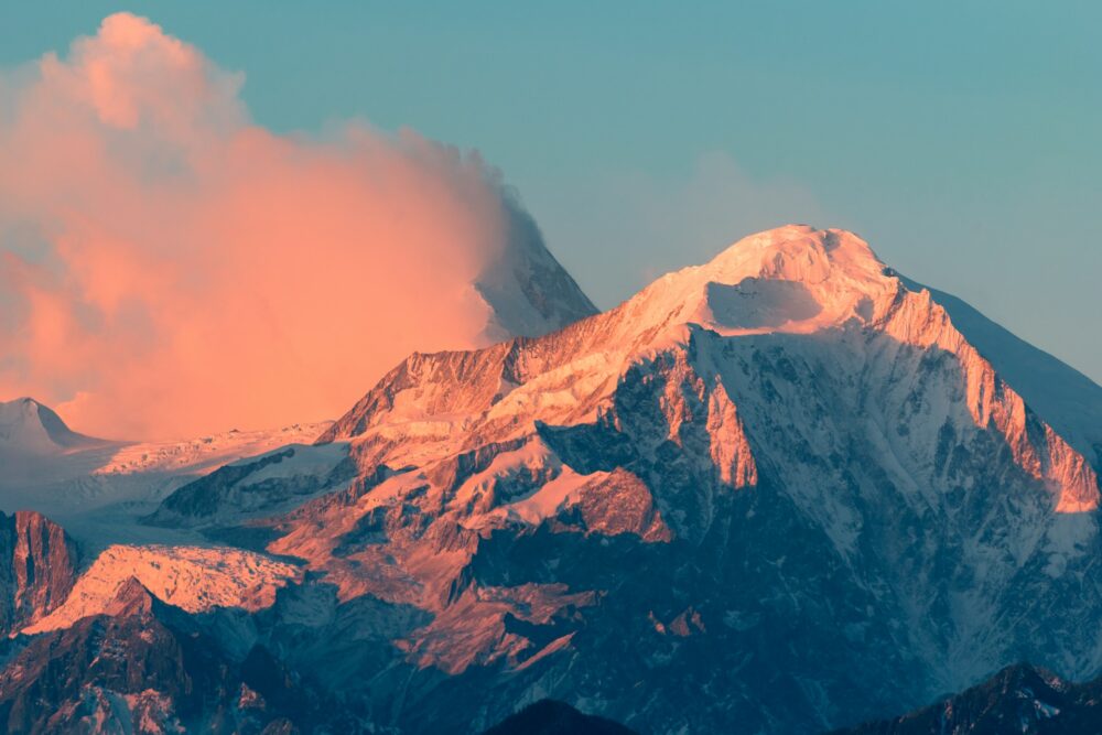 Pink sunlight reflecting on a large snow-covered mountain top and a nearby cloud. The rest of the image is against a clear blue sky.
