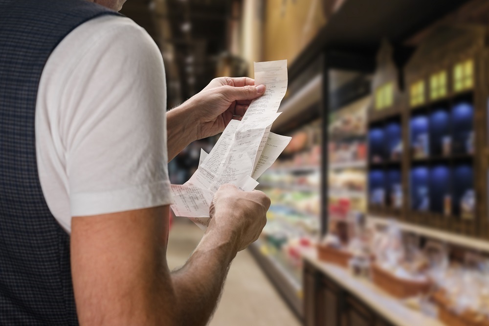 Rear view of a man, wearing a white t-shirt and a dark waistcoat, standing in a shop checking a long receipt.