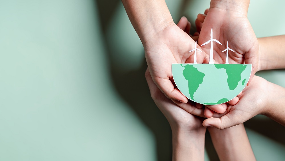 Three sets of hands are all holding a semi circular depiction of the Earth. On top of the semi-circle is a model of 3 small wind farm windmills. The whole image is set against a plain green background.