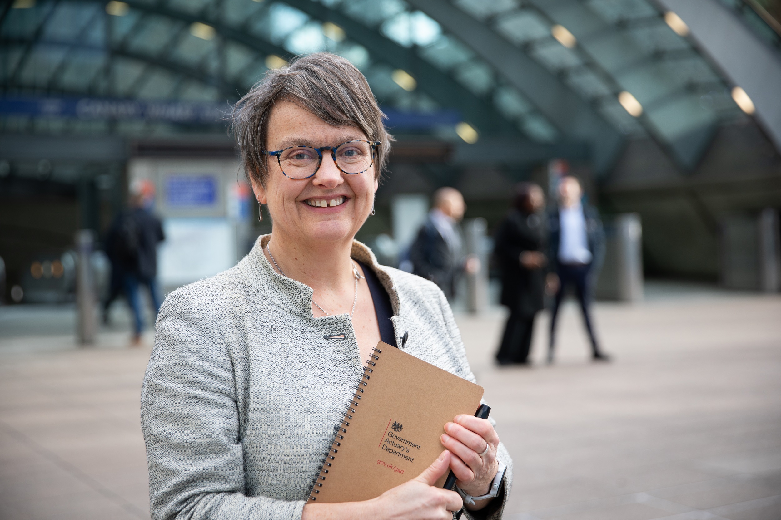 Fiona Dunsire, the Government Actuary holds a notebook with the Government Actuary's Department logo on it. Fiona is outside a London Underground station.