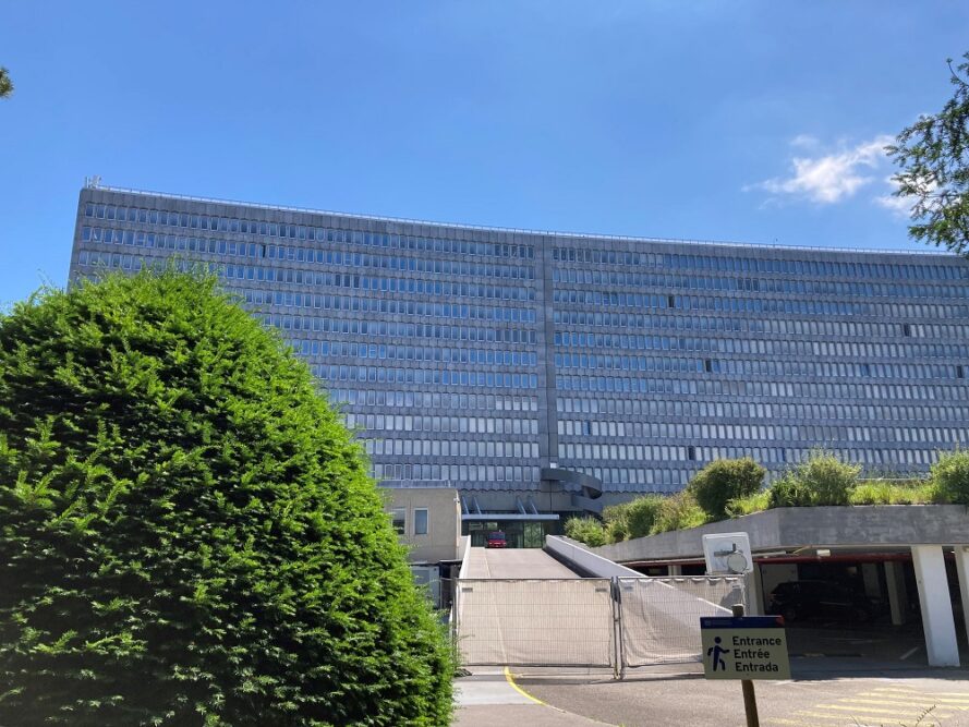 Large office building of grey concrete and hundreds of windows over 11 floors, is pictured against a bight blue sky and with a large green bush in the foreground.