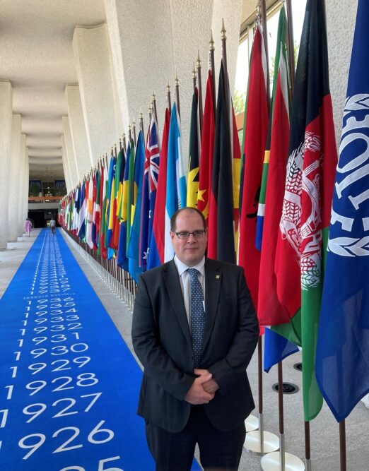 Man in a suit faces the camera standing on a blue carpet on which there are the numbers of years stretching into the distance of the long corridor. To the right of the man there is a row of colourful national flags on flagpoles, stretching into the distance down the log corridor.