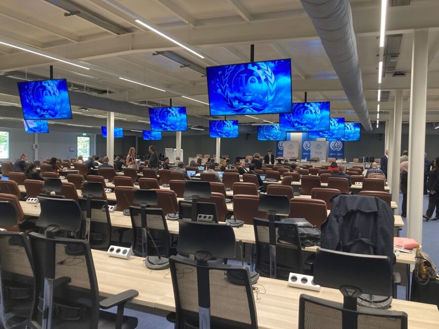 Rear view of large room with rows of tables and chairs, Above, there are several large TV monitors suspended from the ceiling, all displaying the same blue logo.