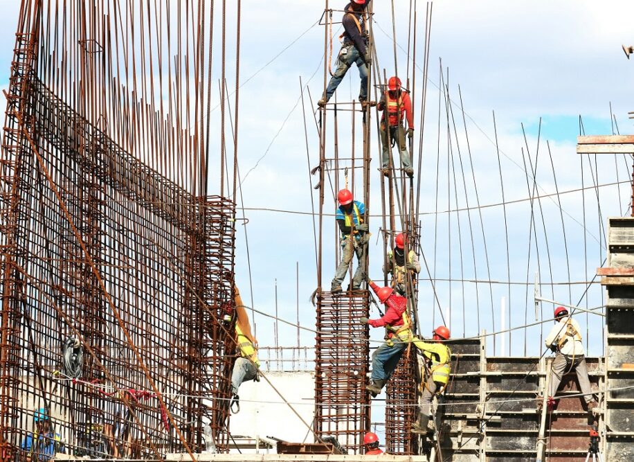 Construction workers wearing red hard hats climbing iron scaffolding on a building site.