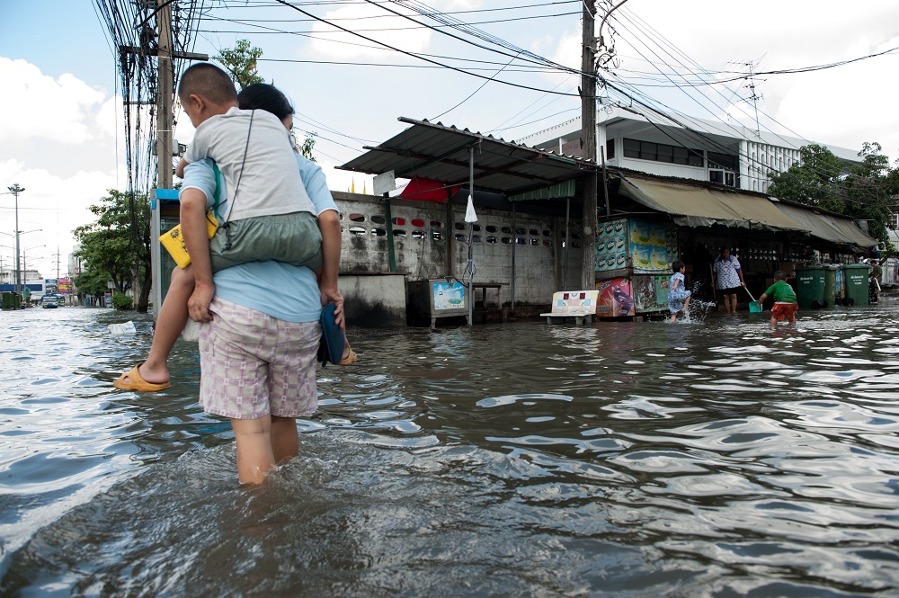 Adult is pictured carrying a child through flood waters in a street. In the background 3 more people are standing in the floodwaters. Above the buildings there are telegraph poles carrying many wires.