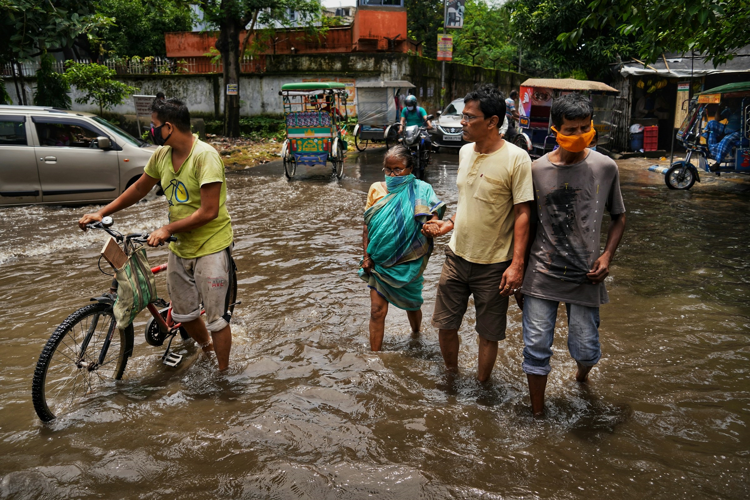 Three men, one with a bike, and an elderly woman, stand in brown rushing flood water in a street. Behind them are tuk-tuks, and cars. Credit: Dibakar Roy Unsplash.