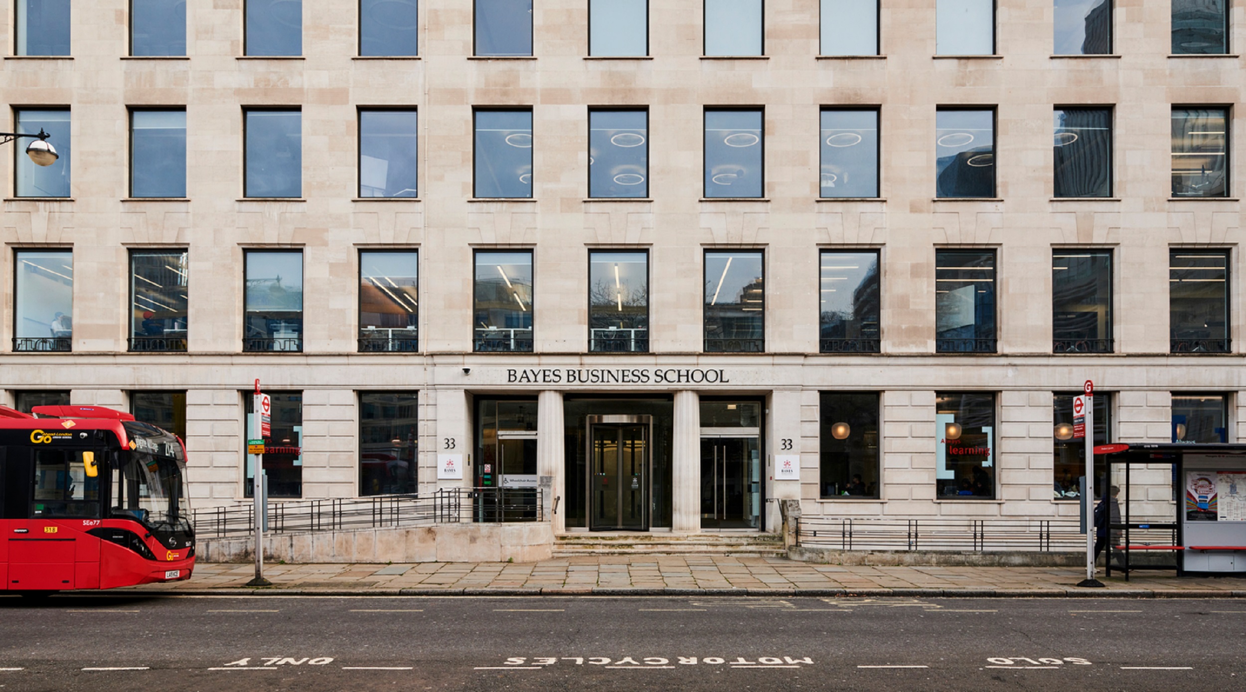 Exterior of Bayes Business School. The building is made of light coloured stone with rows of rectangular windows. There is a red bus on the road in front of the building, near a bus stop.