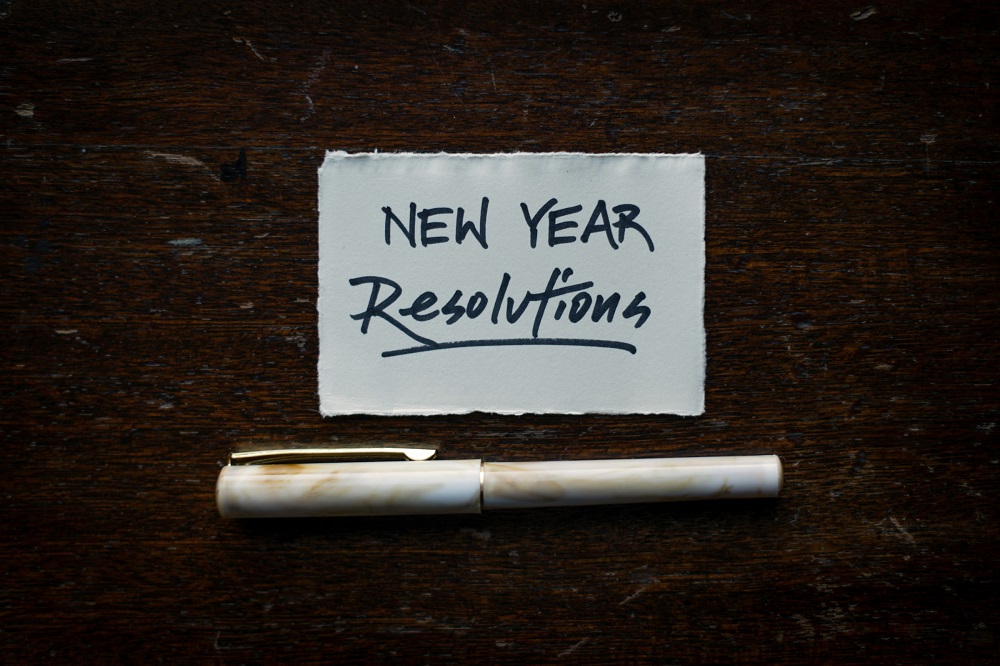 The words 'New Year Resolutions' is written on a small rectangle of white paper placed on a dark surface. Below the white paper is a fountain pen. Credit: Tim Mossholder, Unsplash.