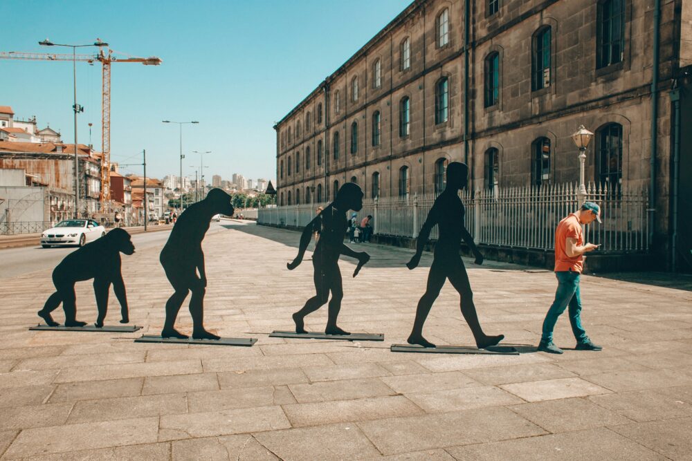 Four black life-sized statues are in a street paved area. They depict the evolution of man, ranging from an ape to a modern human. Next to them is a man wearing jeans and a t-shirt, with his head down checking his mobile phone. Picture credit: Eugene Zhyvchik, Unsplash