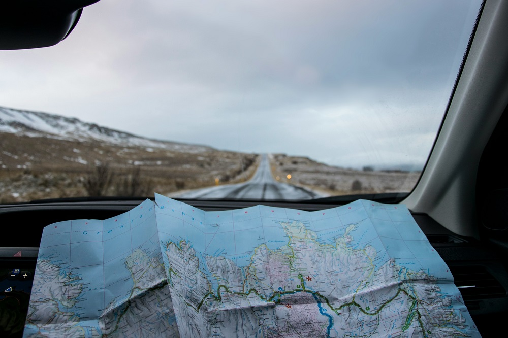 Map of Iceland pictured on dashboard of car. Out of the window is a road going over a wintry landscape, and disappearing over the horizon. Credit: Tabea Schimpf, Unsplash.