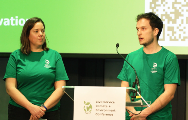 A woman and a man, each wearing green t-shirts, stand at a clear glass podium. On the podium is the sign 'Civil Service Climate + Environment Conference.'