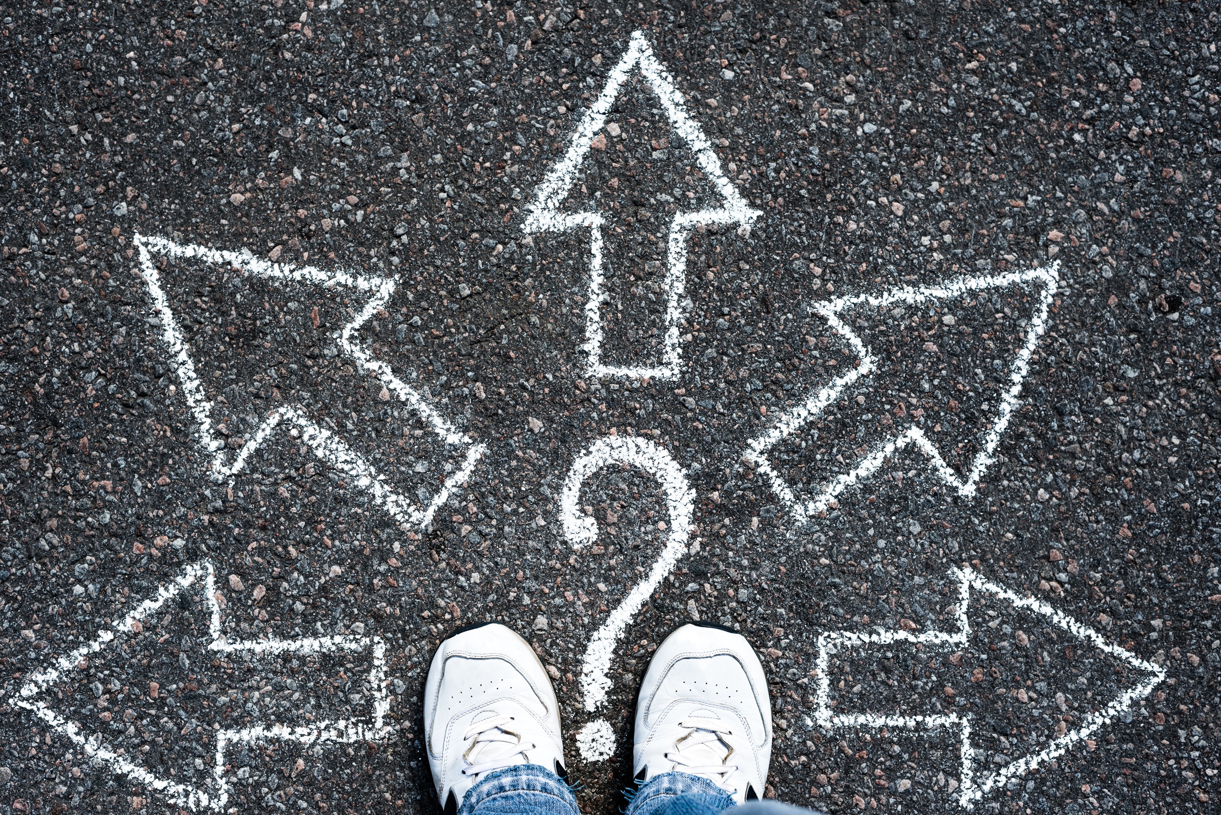 View from above of white trainers on black tarmac on which is drawn 5 arrows and a question mark in white chalk. Credit: Shutterstock