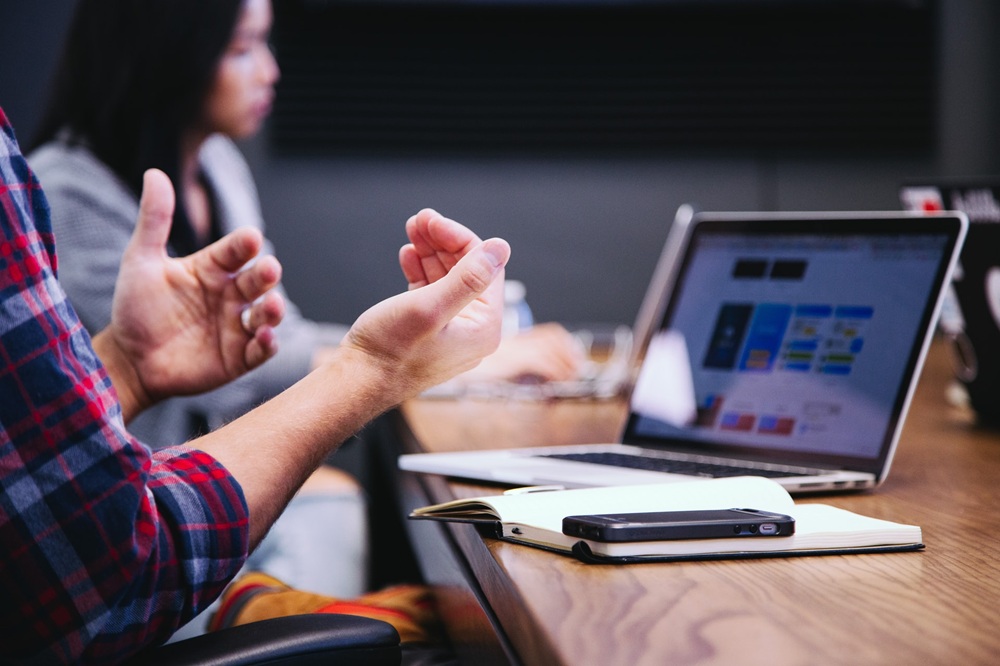 Two people sitting at a wooden table. One is gesturing in front of a laptop. The second is looking forward. Credit: Unsplash.