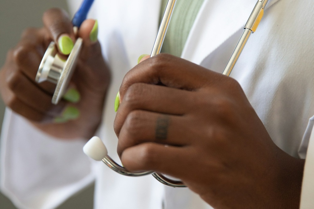 Hands of a black doctor wearing a white coat and sporting bright green nail varnish. The doctor is holding onto a stethoscope. Credit: Nappy, Unsplash.