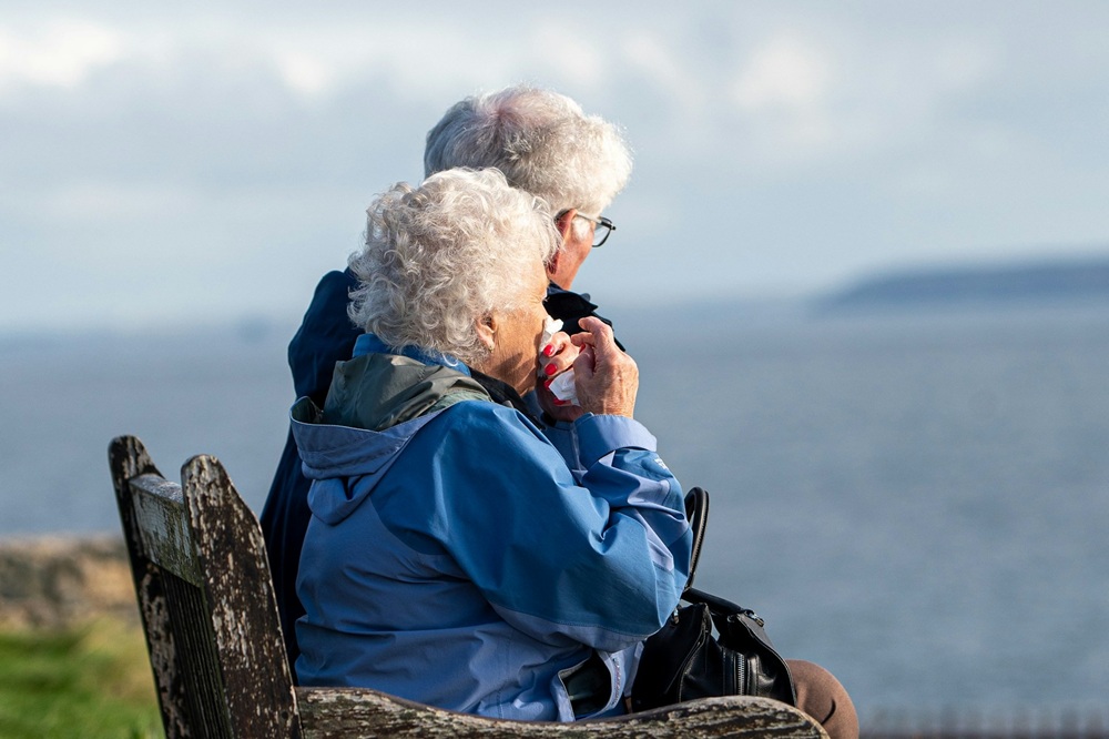 Elderly white-haired couple sitting on an old wooden bench looking out to sea. Credit: Simon Godfrey, Unsplash