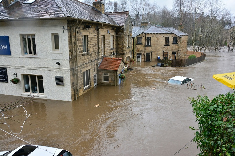 Brown river water flooding outside a pub. A white car parked outside, is virtually submerged in the floodwater. Credit: Unsplash